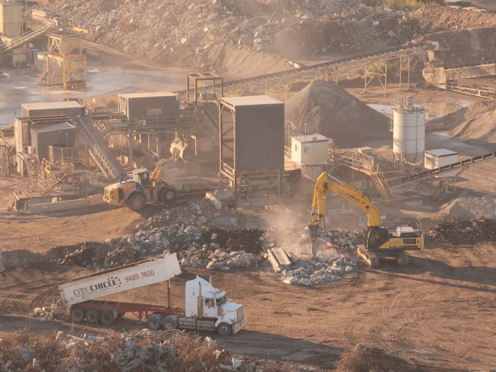 An aerial view of a construction site with heavy machinery, including excavators and trucks, processing material amidst piles of rubble and construction debris.