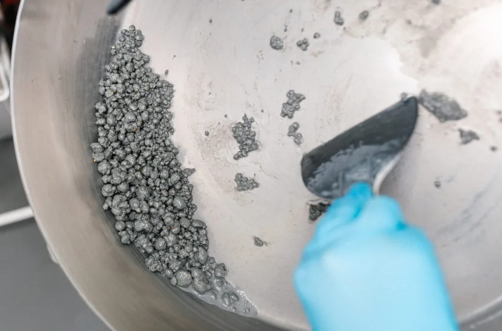 Close-up of a metal mixing bowl with gray, textured material resembling small pellets, with a hand wearing a blue glove using a spatula to scrape the mixture.